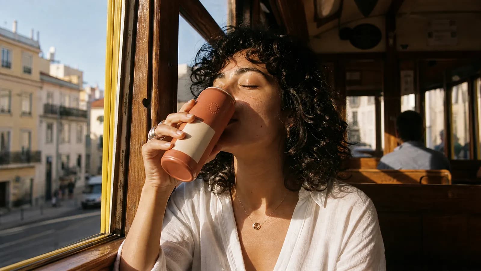 A woman with curly hair sipping from the terracotta can by the window of a yellow Lisbon tram